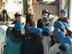 A group of people wearing blue caps listens to a man in glasses standing in front of them, with some flowers and boxes nearby.