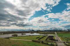 A vast parkland with a body of water, manicured lawns, and industrial structures in the background under a dramatic sky.