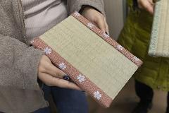 Hands hold a small, decorated tatami mat with a textured, woven surface and a patterned border.