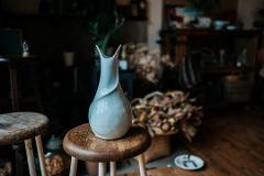 Pottery & Porcelain: Stories of the Artisans Behind the Craft A pale blue, elegantly twisted ceramic vase sits on a rustic wooden stool in a dimly lit studio, surrounded by blurred elements hinting at a craftsperson's workspace.
