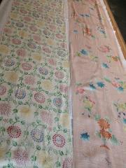 Experience Nagoya "Yuzen" Scarf Dyeing Two bolts of fabric featuring traditional Japanese floral patterns are laid out on a wooden surface, one with a cream base and the other a pale pink.