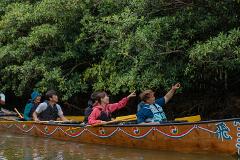 A group of people are in a long, wooden boat with paddles, surrounded by lush green trees, and two people in the boat are pointing upwards.
