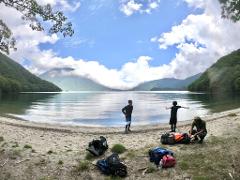 Hikers with backpacks prepare for an adventure on the shore of a serene lake with a majestic mountain in the background.