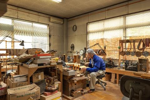 An elderly craftsman sits in his workshop, surrounded by tools and materials, meticulously working on a comb.