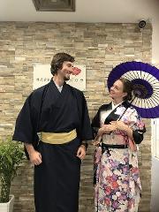 A smiling couple dressed in traditional kimonos stands in front of a stone wall, holding a purple umbrella.