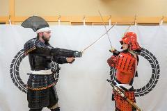 Two people dressed in samurai armor face each other, holding swords in a staged combat pose against a white backdrop.