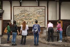 A group of people admire a large tiled artwork depicting a traditional Japanese scene outside a historical building.