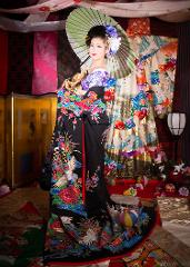 Eine Frau in einem schwarzen Kimono mit buntem Kranich- und Blumenmuster posiert mit einem grünen Sonnenschirm inmitten einer kunstvollen japanischen Kulisse mit Stoffbahnen und Requisiten.
