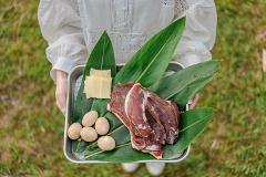 A person in a white dress holds a tray with meat, cheese, and eggs on green leaves.