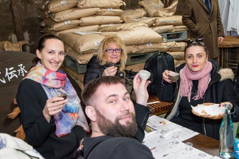 Three people are enjoying a sake tasting in front of a wall of sake bags.