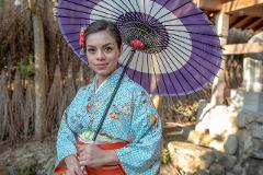 A woman in a blue kimono with orange accents holds a purple and white umbrella while standing in a garden.