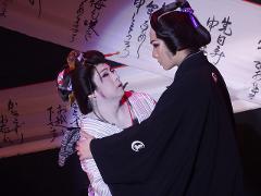 Two Kabuki actors, dressed in traditional garb and makeup, are locked in an intense, emotional embrace on stage, with Japanese calligraphy in the background.