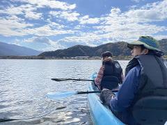 Two people in a blue kayak paddle on a lake with mountains in the background under a cloudy sky.