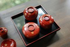 Discover the Art of Yamanaka Lacquerware & Experience the Ultimate Hospitality of the Tea Ceremony Three lidded red lacquerware bowls decorated with gold flowers sit on a black tray.