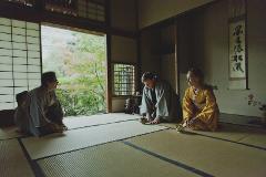 Three people in traditional Japanese clothing are kneeling on tatami mats in a room with a view of greenery outside.