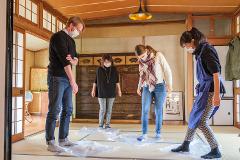 Four people wearing face masks stand in a room, each stepping on a clear plastic sheet that is placed on a tatami mat floor.