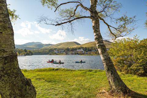 People kayak on a tranquil lake surrounded by green hills, trees, and buildings under a clear blue sky.
