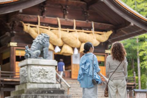 Nagano 2-Day Suwa Shrine Tour & Lake Onsen Stay with Sakura Two women stand at the base of stairs leading to a shrine, with a stone lion statue in the foreground and a traditional Japanese building in the background.