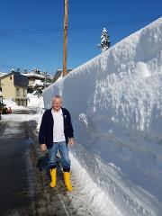 Professional breeder-guided Koi farm tour in the sanctuary of Nishikigoi A man in yellow boots stands beside a massive snow wall under a bright blue sky, with snow-covered trees and houses visible in the background.