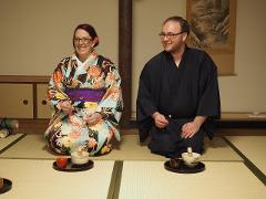 A man and woman in traditional Japanese clothing kneel on tatami mats preparing tea.