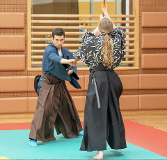 Dvaja ľudia v tradičnom japonskom oblečení cvičia s drevenými mečmi v prostredí podobnom dojo.