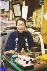 An elderly Japanese woman with distinctive eye makeup smiles warmly while wearing a traditional indigo jacket with white lettering, surrounded by shelves of papers and craft supplies.