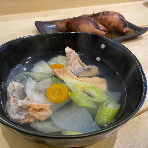 This image showcases a bowl of Sanpei soup, featuring salmon, radish, carrots, and leeks, complemented by a plate of grilled squid in the background.