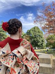 A person in a red kimono and elaborate obi stands in a park with a blue sky and autumn trees.