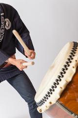 A person in traditional Japanese attire plays a large drum with two wooden sticks.