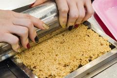Hands with glittery nail polish press a metallic roller over a pan of puffed rice cereal to make a Japanese snack.