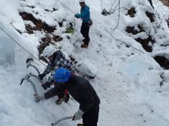 Three people are ice climbing up a snow-covered slope, using ice axes and wearing helmets.