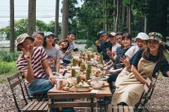 A diverse group of people smile and pose for a photo around a table laden with food at an outdoor festival.