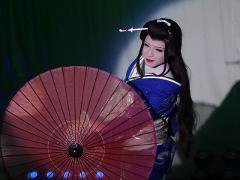A person in a Japanese theatrical costume with full makeup holds a red umbrella, looking to the side.