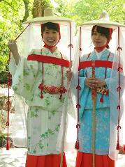Two women in traditional Japanese attire, featuring wide-brimmed hats with sheer veils, stand amidst lush greenery, one smiling and the other holding a staff.