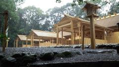 A Japanese shrine with wooden structures and thatched roofs is set against a backdrop of lush green trees under a cloudy sky.