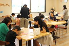 Plusieurs enfants sont assis à des tables dans une salle lumineuse, participant à une activité avec l&#39;aide d&#39;adultes.