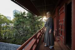 Meisatsu Zojoji Temple - A Journey to Tokyo’s Cultural Origins A man in traditional Japanese attire stands on a wooden balcony overlooking trees and city buildings.