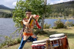 A man in traditional clothing plays a conch shell by a lake with drums nearby.