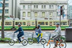 Three people on bicycles ride alongside a green and beige tram on a city street in Japan.
