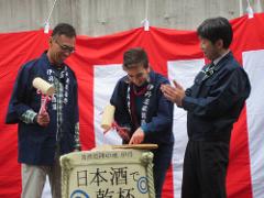 Three people are participating in a Japanese ceremony, with a banner of red and white stripes behind them. One person is about to strike a wooden plank atop a sign, while the other two people watch and clap. The sign has Japanese writing on it.