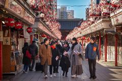 A group of people walk down a bustling street lined with shops and decorated with red and white lanterns, under a clear blue sky.