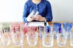 A person in a blue cardigan holds a clear glass in front of a table filled with various colorful Edo Kiriko glasses.