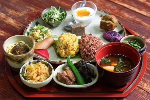This vibrant image shows a beautifully arranged spread of traditional Okinawan cuisine, featuring colorful rice balls, miso soup, and various side dishes on an ornate platter.