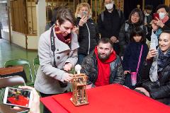 A group of people, some wearing masks, gather around a table with a wooden mechanical doll and a woman holding up a phone to take a photo.