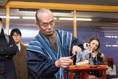 An elderly man with a mustache demonstrates the operation of a mechanical doll in front of a crowd.