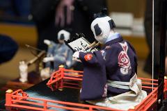 A detailed close-up shot shows a Japanese doll in traditional attire holding brushes to write calligraphy.