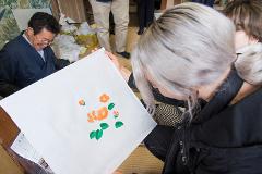 A man in a dark blue robe shows a white cloth with orange flowers and green leaves to a woman with light grey hair.