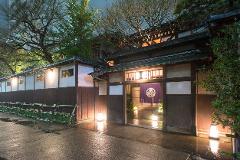A traditional Japanese restaurant entrance glows with warm lights on a wet evening, featuring bamboo fencing and an entrance curtain with a family crest.