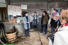 Tourists observe a traditional sake brewery's water source, with a guide explaining the process.