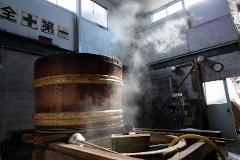 A large wooden barrel steams in a rustic brewery, hinting at the traditional process of sake making.